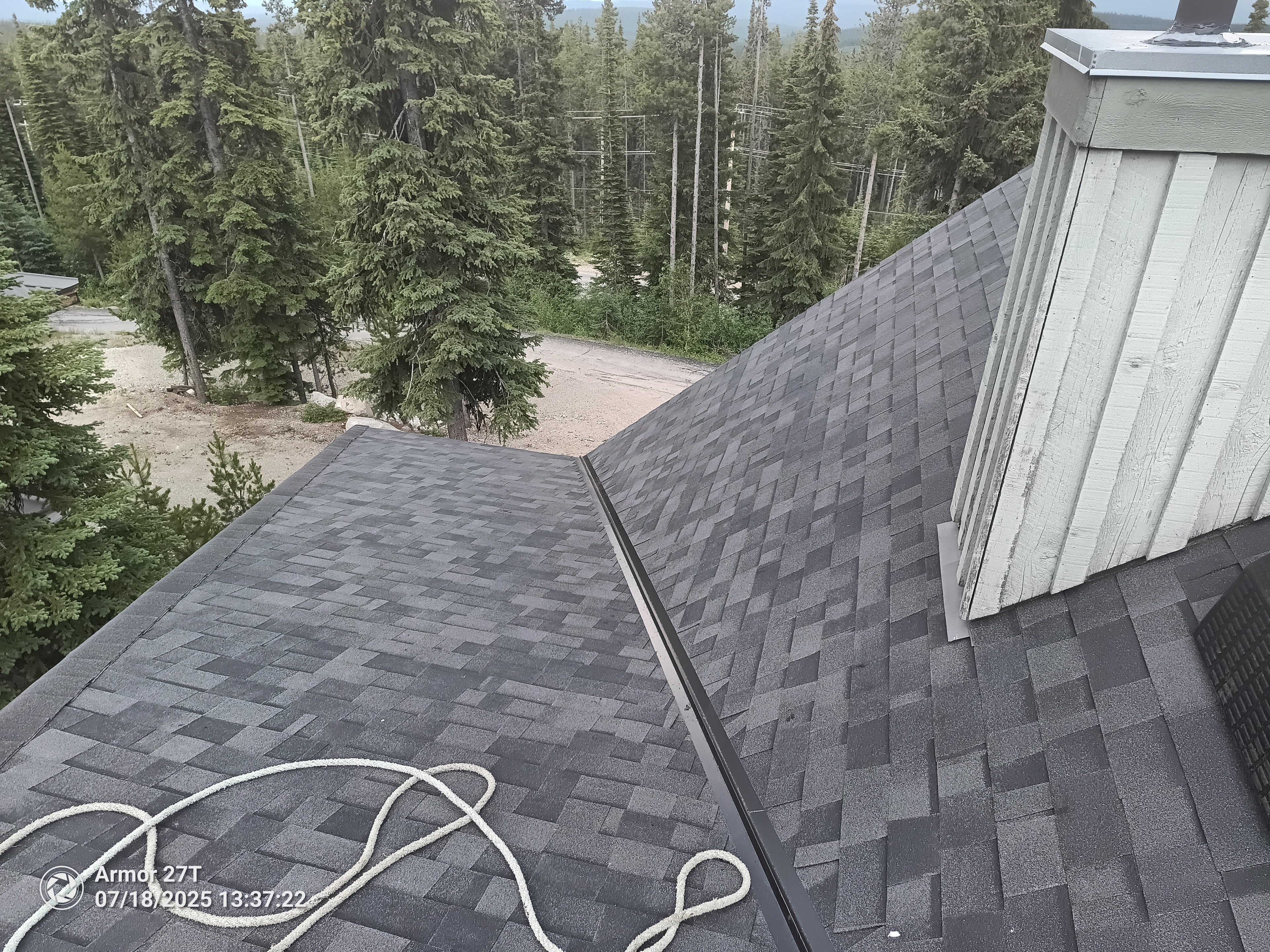 Completed dark shingle roof with ridge cap and forest backdrop